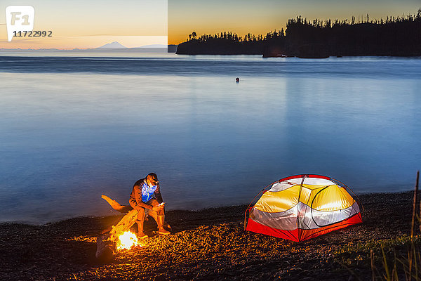 Ein Lagerfeuer am Strand bei Sonnenuntergang beleuchtet ein nahegelegenes Zelt. Ein Mann sitzt am Feuer und schaut auf sein Handy  während das ruhige Meerwasser das warme Licht im Hintergrund reflektiert; Hesketh Island  Homer  Alaska  Vereinigte Staaten von Amerika'.