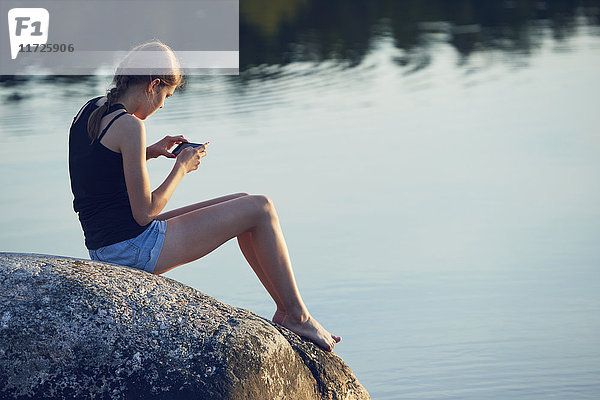 Teenager-Mädchen sitzt am See und benutzt ein Handy
