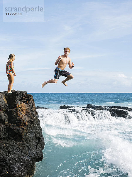 Mädchen (6-7) steht auf einer Klippe und ein Mann springt ins Meer