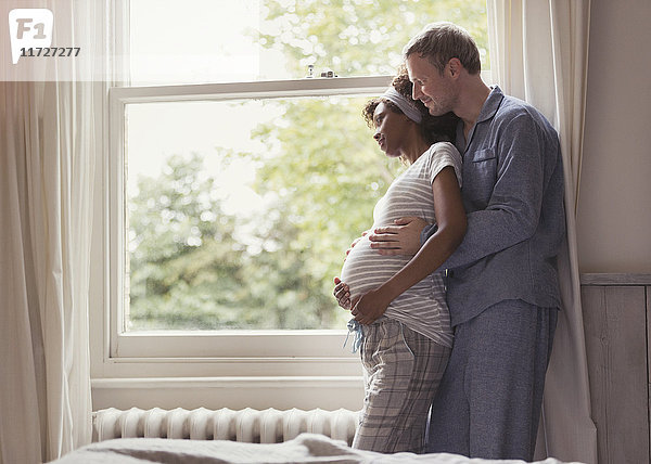 Zärtliches schwangeres Paar im Pyjama  das sich am Fenster umarmt