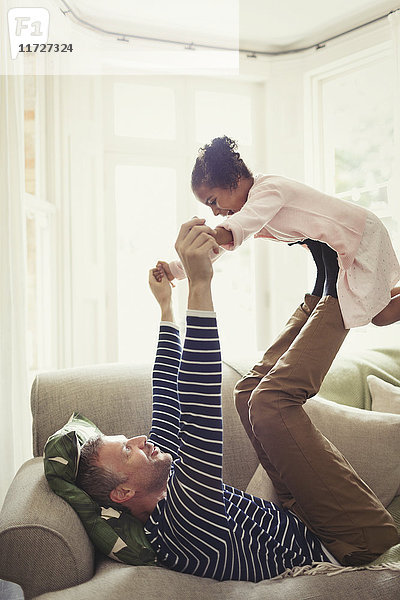 Multiethnischer Vater beim Spielen  balancierende Tochter auf Beinen über Kopf auf dem Sofa