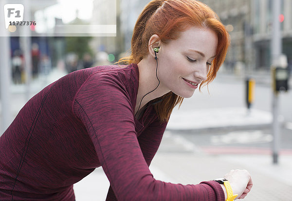 Lächelnde Läuferin mit roten Haaren und Kopfhörern  die auf intelligente Uhr achtet