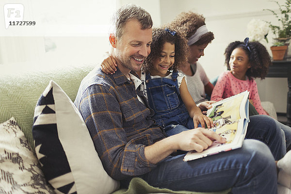Lächelnder multiethnischer Vater liest Buch mit Tochter auf Sofa