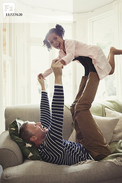 Verspielter multiethnischer Vater  der die Tochter auf den Beinen auf dem Sofa balanciert.