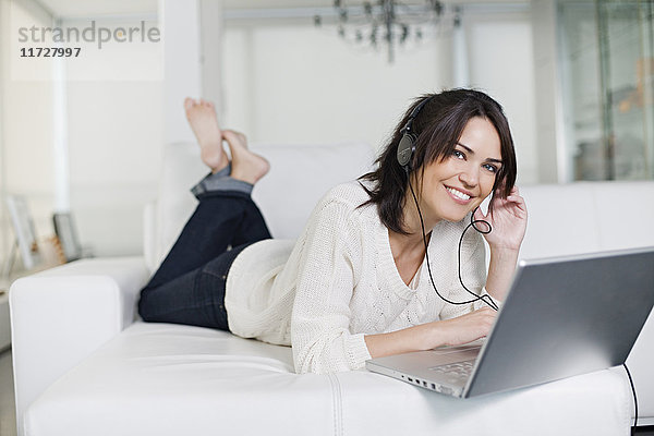 Schöne brünette Frau auf einer Couch mit Computer und Kopfhörer lächelnd vor der Kamera