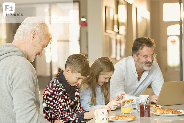 Männliche schwule Eltern und Kinder beim Frühstücken und mit Laptop und digitalem Tablett am Küchenarbeitsplatz