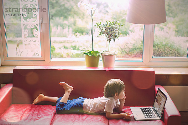 Junge mit Laptop auf rotem Ledersofa im Wohnzimmer