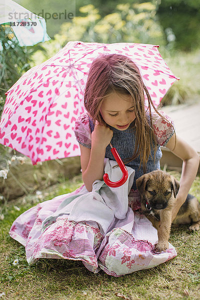 Mädchen mit Hundewelpe hält herzförmigen Regenschirm im Gras