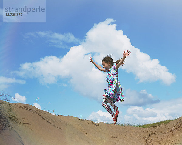 Überschwängliches Mädchen springt vor Freude am Strand Hügel unter sonnigen blauen Himmel mit Wolken