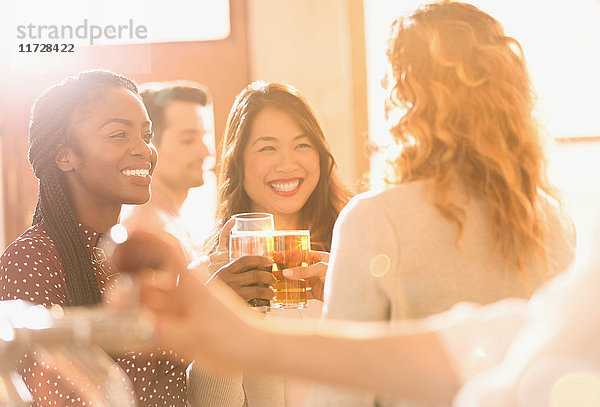 Lächelnde Freundinnen stoßen mit Biergläsern in einer sonnigen Bar an