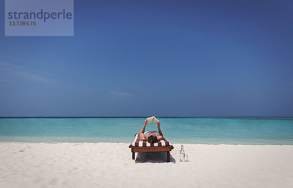 Frau entspannt sich  liegt und liest ein Buch auf einem Liegestuhl am sonnigen tropischen Strand