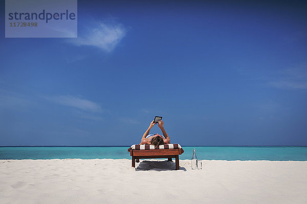 Frau entspannt sich  nimmt ein Sonnenbad  liegt auf einem Liegestuhl und benutzt ein digitales Tablet am sonnigen tropischen Strand