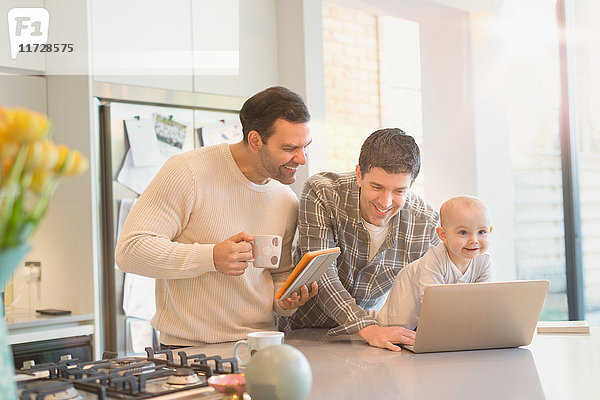 Männliche schwule Eltern mit Baby-Sohn mit digitalem Tablett und Laptop in der Küche