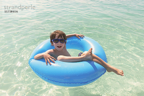 Porträt lächelnder Junge mit Sonnenbrille schwimmt in blauem aufblasbarem Ring im Meer