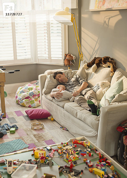 Erschöpfter Vater und Sohn schlafen auf dem Sofa im unordentlichen Wohnzimmer mit Spielzeug.