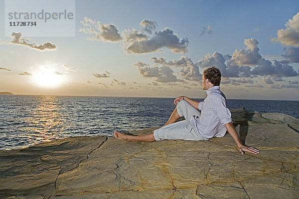 Rückansicht eines Mannes  der den Sonnenuntergang in einer Klippe am Meer betrachtet.