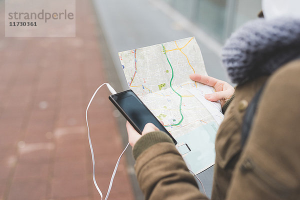 Über-Schulter-Ansicht einer Rucksacktouristin mit Smartphone beim Blick auf die Karte auf dem städtischen Bürgersteig