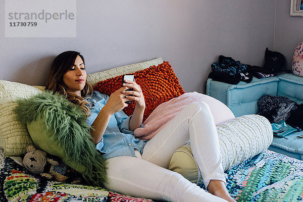 Junge Frau liegt auf schäbig-schickem Bett und schaut auf Smartphone