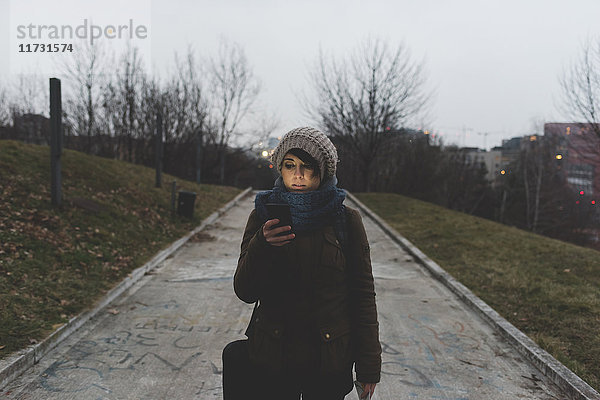 Backpackerin geht in der Abenddämmerung im Stadtpark spazieren und schaut auf ihr Smartphone