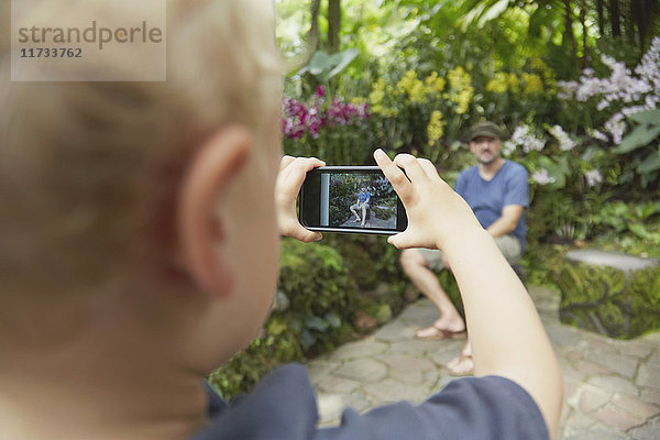 Über-die-Schulter-Ansicht eines Jungen  der seinen Vater mit einem Smartphone im Botanischen Garten fotografiert  Singapur