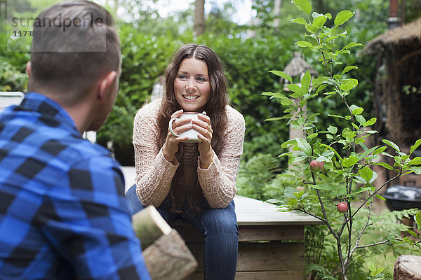 Junge Frau trinkt Kaffee auf der Veranda eines Chalets  während ihr Freund Baumstämme trägt