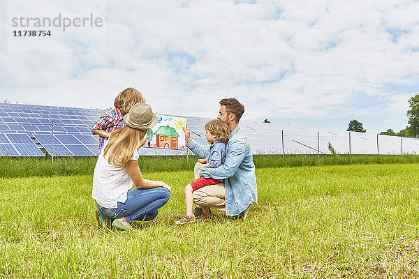Junge Familie sitzt auf dem Feld und schaut auf eine Kinderzeichnung von einem Haus neben einem Solarpark