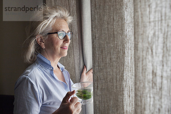 Ältere Frau hält Getränk in der Hand und schaut aus dem Fenster