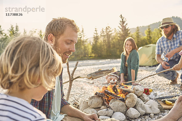 Familie sitzt am Lagerfeuer und kocht Fisch an einem Ast