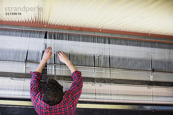 Draufsicht eines jungen männlichen Webers  der Fäden auf einer alten Webmaschine in einer Textilfabrik einstellt