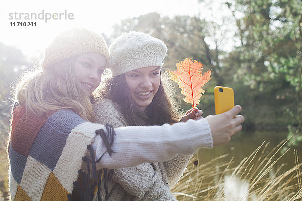 Teenager-Mädchen in Strickwaren am Fluss halten Herbstblatt und benutzen Smartphone  um sich selbst zum Lächeln zu bringen