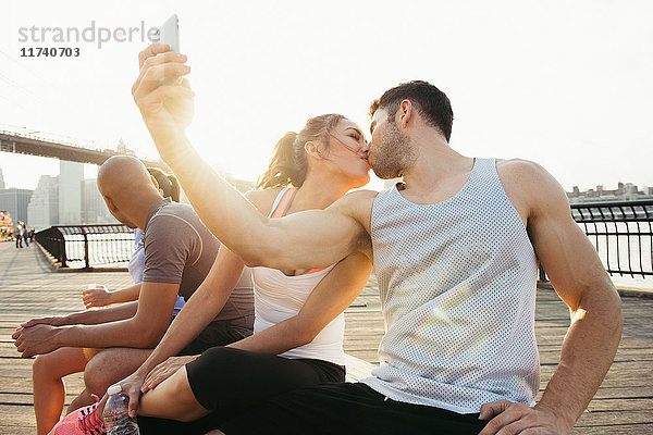 Junger Erwachsener  laufendes Paar küsst sich für Smartphone-Selfie am Flussufer  New York  USA