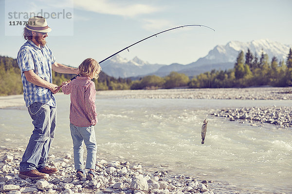 Mittelgroßer erwachsener Mann und Junge in Flussnähe  die eine Angelrute mit Fisch in der Hand halten  Wallgau  Bayern  Deutschland