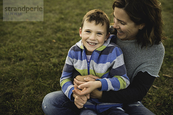 Junge sitzt auf dem Schoß der Mutter im Gras und schaut lächelnd in die Kamera