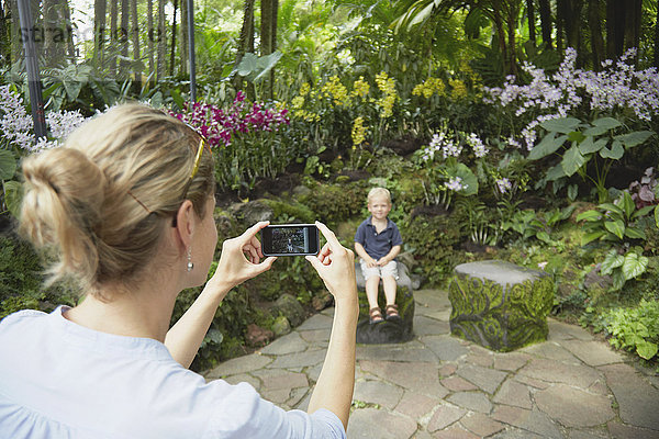 Blick über die Schulter einer Frau  die ihren Sohn auf einem Smartphone im Botanischen Garten in Singapur fotografiert
