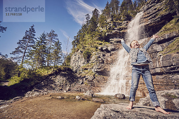 Junges Mädchen steht auf einem Felsen neben einem Wasserfall  die Arme aufgeregt erhoben