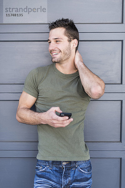 Porträt eines jungen Mannes mit Smartphone  der sich an eine graue Tür lehnt
