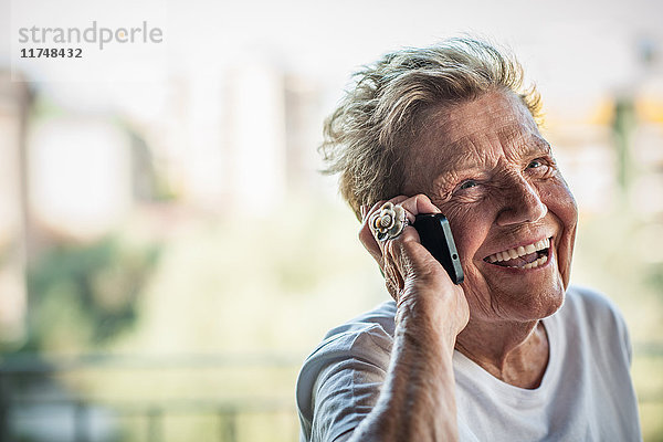 Eine hochbetagte Frau plaudert und lacht am Smartphone auf dem Balkon der Wohnung