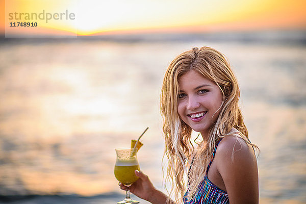 Porträt einer jungen Frau  die bei Sonnenuntergang am Strand einen Cocktail trinkt