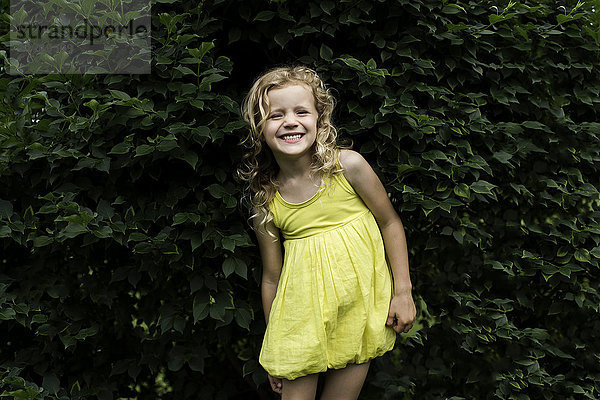 Bildnis eines lächelnden blonden Mädchens in gelbem Kleid vor einer Gartenhecke stehend