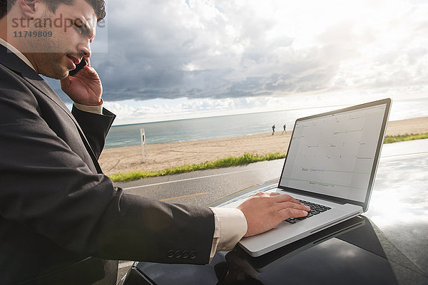 Geschäftsmann spricht mit einem Smartphone  während er sein Laptop auf einem Parkplatz an der Küste benutzt