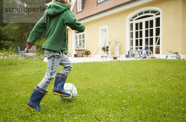 Rückansicht eines Jungen mit Gummistiefeln beim Fussballspielen im Garten