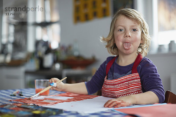 Porträt eines Jungen  der am Tisch sitzt  einen Pinsel hält und die Zunge herausstreckt