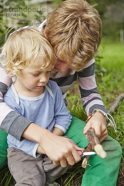 Junge sitzt mit seinem jüngeren Bruder mit einem Taschenmesser auf einem Zweig im Garten
