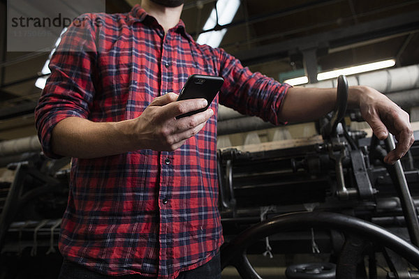 Ausschnitt einer Aufnahme eines männlichen Weber-Smartphones neben einer alten Webmaschine in einer Textilfabrik