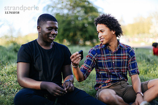 Zwei männliche Freunde sitzen im Gras und schauen auf ein Smartphone