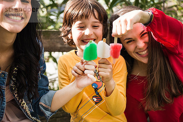 Zwei junge Frauen und ein Junge sitzen auf einer Bank und halten Eislollies zusammen  um die Farben der italienischen Flagge zu kreieren