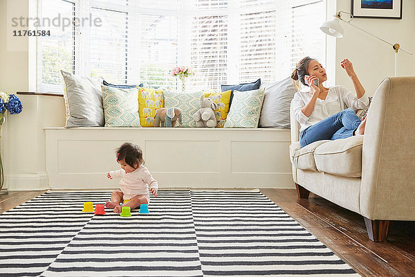 Frau mit Baby-Tochter auf Sofa liegend beim Smartphone-Anruf