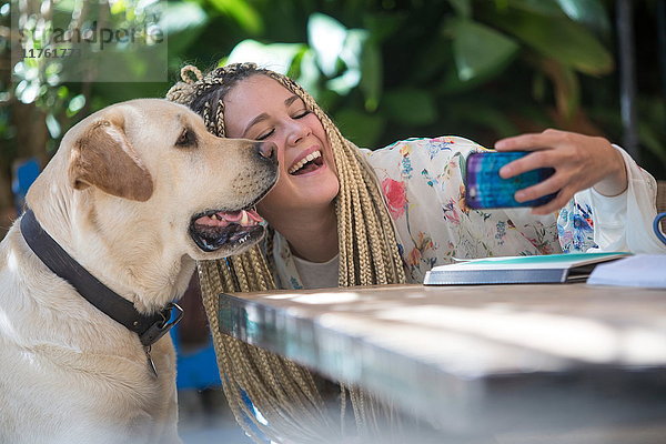 Junge Frau mit Dreadlocks  sitzt im Freien  nimmt Selfie mit Hund  benutzt Smartphone