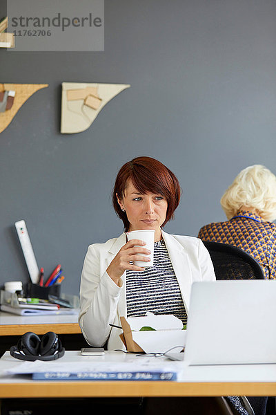 Designerin schaut auf Laptop und trinkt Kaffee am Bürotisch