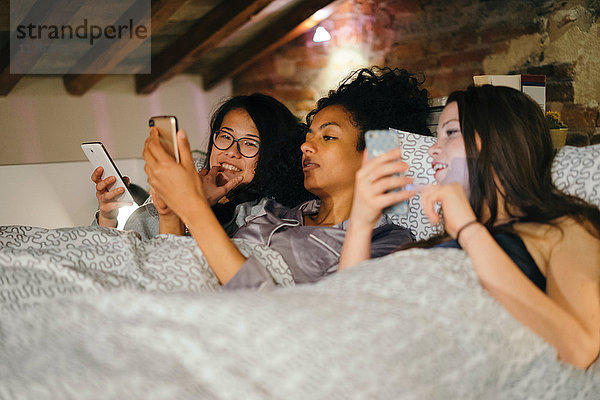 Freunde liegen nebeneinander im Bett und schauen auf Mobiltelefone